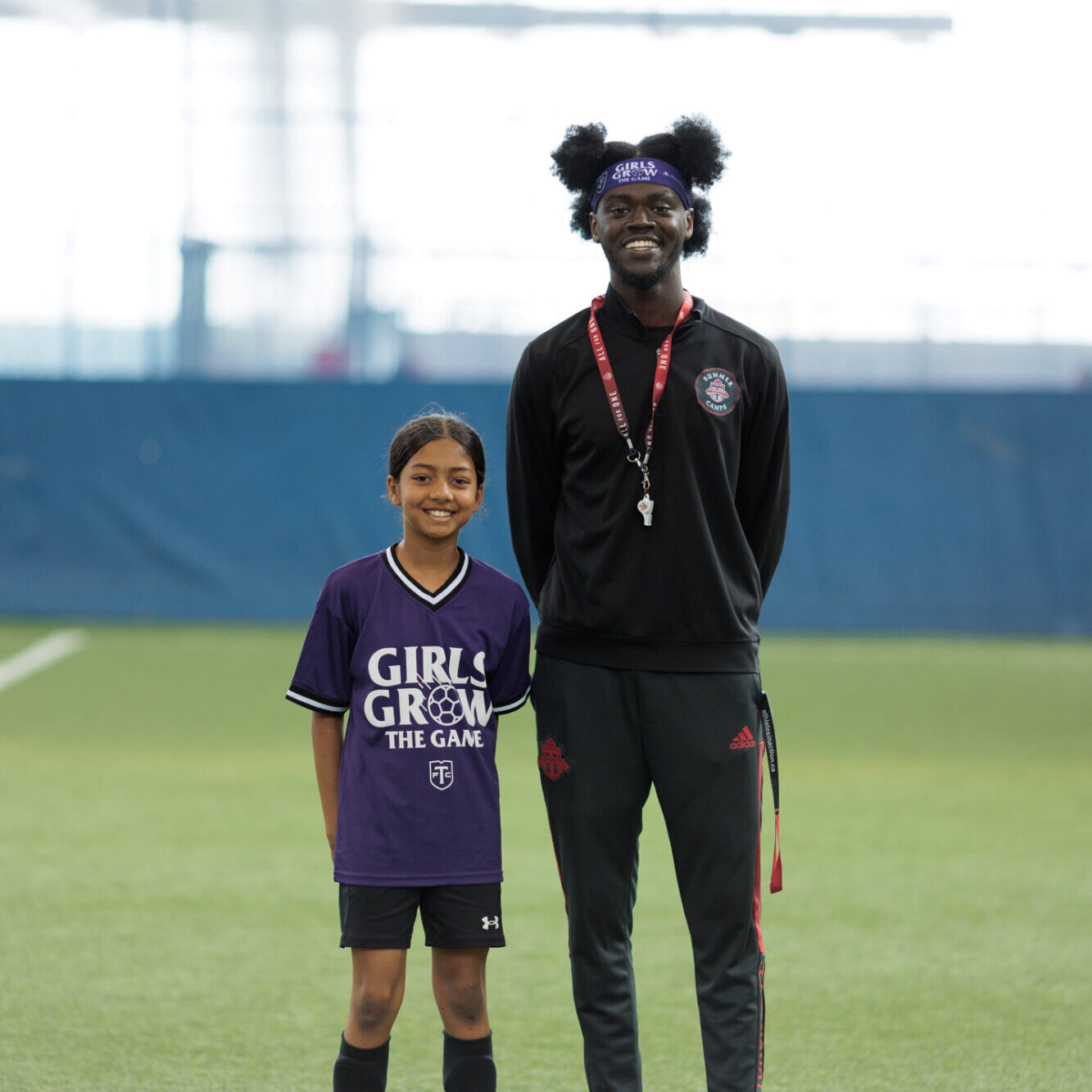 A soccer coach in a black jacket and black pants stands next to a young player wearing a purple jersey and black shorts.