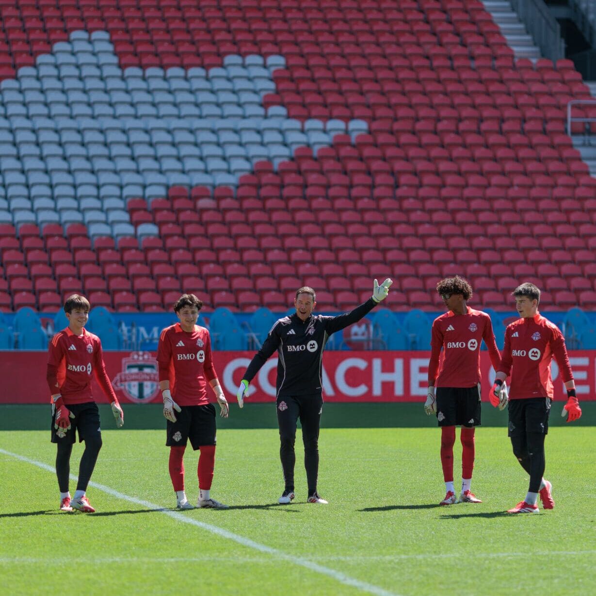 TFC_COH_25-62001 Soccer coach in all black stands in between four young soccer players wearing red jerseys and black shorts on a soccer field with stadium seating in the background.